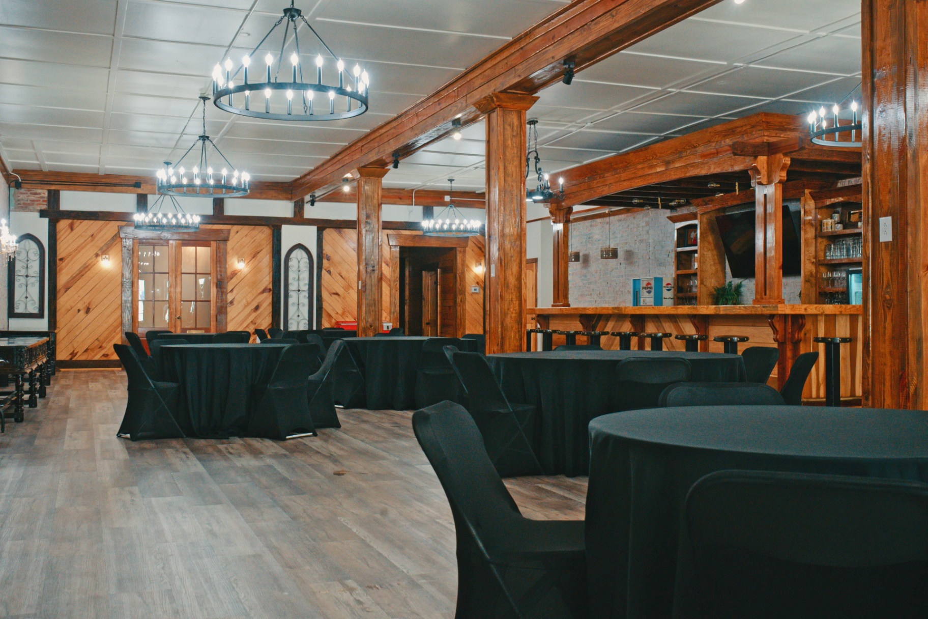 The downstairs event venue set with round tables, timber beams, and wagon-wheel chandeliers